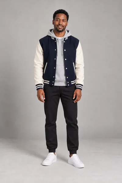 Homem de jaqueta college azul-marinho e creme sobre moletom cinza com capuz, calça jogger preta e tênis branco — prévia do trocador de roupa com IA